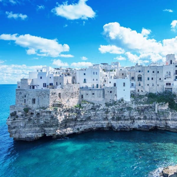 White Italian buildings perched on a cliff overlooking the turquoise Adriatic Sea under a blue sky.