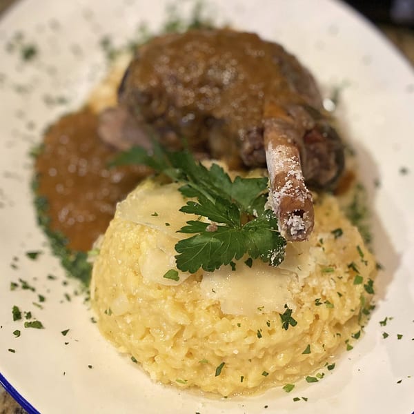 Braised lamb shank with rich sauce, creamy risotto, parmesan shavings, and fresh parsley on a white plate.